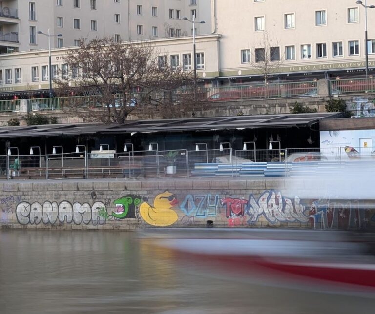 Zwischen Stillstand und Strömung: Während die gelbe Ente mit stoischer Gelassenheit die Wand hütet, zieht das Leben am Donaukanal in Unschärfe vorbei – ein bunter Beleg für die gezähmte Rebellion am Lauf der Zeit.