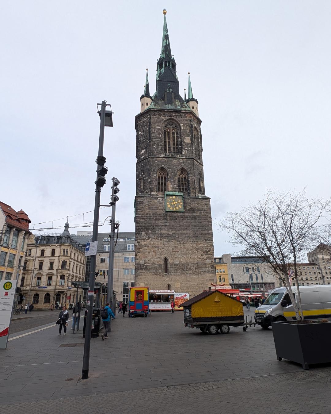 Der historische Rote Turm auf dem Marktplatz in Halle (Saale) mit einer großen Turmuhr unter bewölktem Himmel, im Vordergrund Marktstände und Passanten.