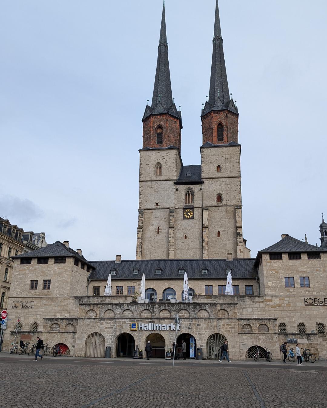 Blick vom Hallmarkt in Halle (Saale) auf die zwei Blauen Türme der Marktkirche Unser Lieben Frauen über einem Sandsteingebäude mit Rundbögen.