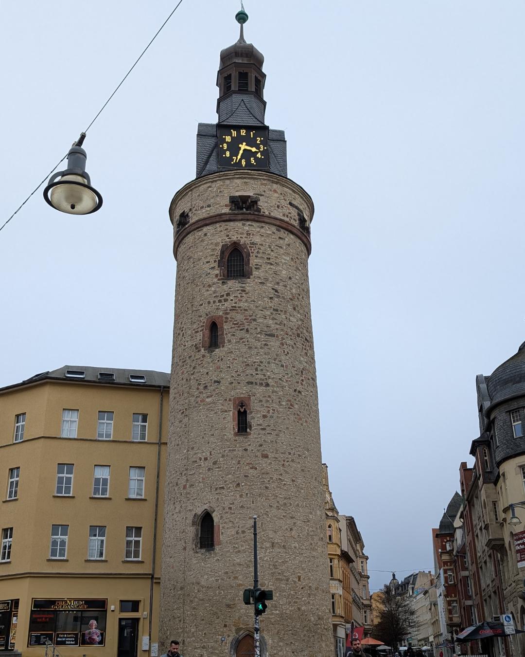 Der historische Leipziger Turm in Halle (Saale), ein runder mittelalterlicher Wartturm aus Bruchstein mit einer Turmuhr an der Spitze, flankiert von Stadthäusern.