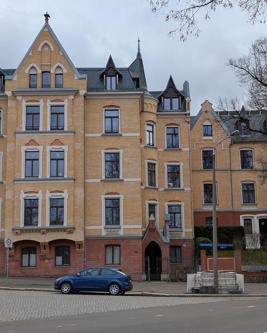 Mehrstöckiges historisches Eckhaus in Halle (Saale) mit gelber Backsteinfassade, rotem Erdgeschoss und markanten neogotischen Giebeln unter grauem Himmel.